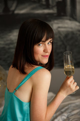 Woman portrait at party. Lovely, beautiful, young girl in gentle silk pajama, holding a glass of champagne, posing while sitting on the bed. Indoors.