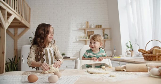Little caucasian girl is learning how to bake, preparing pastry while her younger brother in baby chair is playing and positively smiling - togetherness, childhood dream concept 4k
