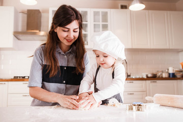 The little daughter in the chef's hat and apron and her mother prepare baking in the bright, classic kitchen.