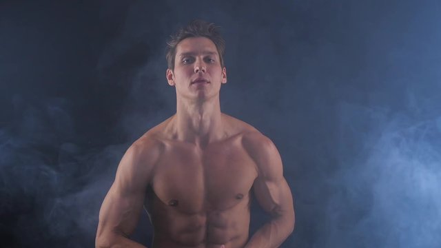 Muscular shirtless man posing on dark background, with smoke around him.