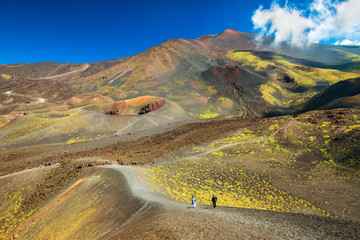Beautiful valley landscape of The Mount Etna, Sicily, Italy. Colorful lava hills covered with plants and grass. People walking along the road between volcanic rocks and craters