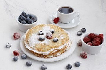 Pancakes sprinkled with powdered sugar, raspberries, blueberries and a cup of coffee on a light background