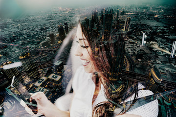 Beautiful smiling Caucasian brunette using smart phone while sitting in car. On picture cityscape. Double exposure technique used.