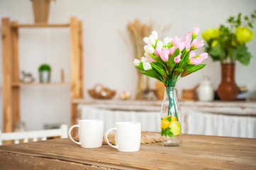 Breakfast cups and fruit. Spring tulips on the table. Wooden table in a bright rustic-style kitchen. Scandinavian style in the interior of the kitchen.