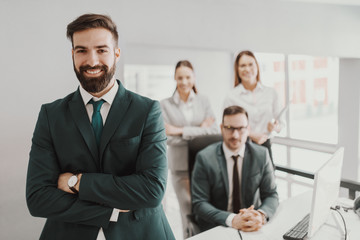 Young handsome bearded smiling CEO in suit standing in office with arms crossed. In background his successful team posing.