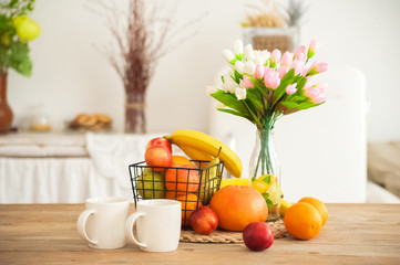 Breakfast cups and fruit. Spring tulips on the table. Wooden table in a bright rustic-style kitchen. Scandinavian style in the interior of the kitchen.