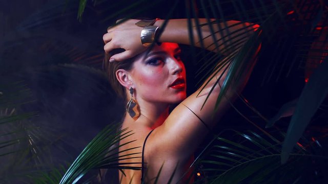 Beautiful girl in luxurious expensive dress, with large stylish gold earrings and bracelet on her hand, stands among tropical leaves. Concept for advertising jewelry. Red light.