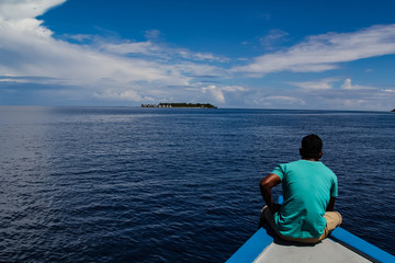 Man sitting on a traditional wooden boat heading over the calm tropical ocean towards a luxury...