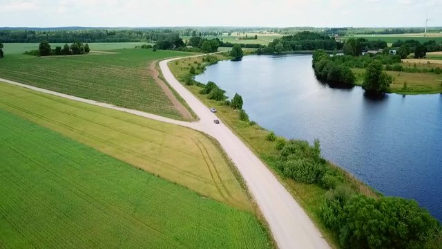 Aerial, tilt up, drone shot following a car driving on the countryside, passing by green grass fields and a blue river, on a cloudy summer day, in Zemaitija region, Lithuania