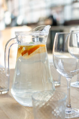 Pitcher of homemade lemonade drink of soda water, lemon, orange and fresh mint leaves on wooden table with glasses foreground. Sunny day blurred background