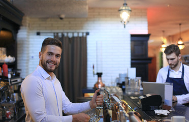 Handsome bartender is smiling and filling a glass with beer while standing at bar counter in pub.