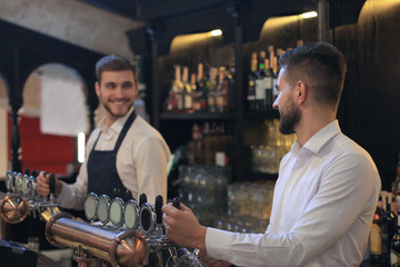 Handsome bartender is smiling and filling a glass with beer while standing at bar counter in pub.