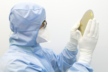 An engineer working in a clear room wearing a special uniform and protective glasses holds silicon wafer with microchips in hands in gloves