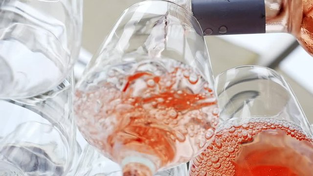 Pouring cold rose wine into glasses. Elegantly lined up row of wine glasses with bottles of alcohol stand on white tablecloth