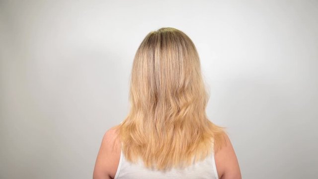 Close up back view of blonde white woman standing still before camera then begining to comb her hair with hairbrush.