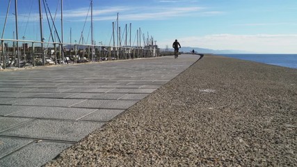 People run on the waterfront. Sunny summer day. Jogging in the fresh sea air. People ride bicycles and take pictures of the sea. Azure birch. Rocky promenade by the sea. Man runs on the street - Powered by Adobe