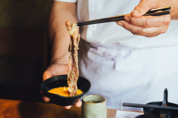 A hand pinching boiled well done slice Wagyu beef with marble texture with chopsticks and dip in...
