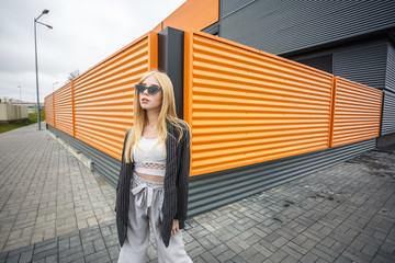 closeup portrait of cute smiling little model girl in striped jacket and sunglasses posing near gray corrugated striped orange gray wall