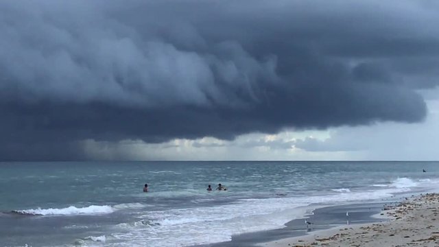 Hurricane with thick ominous clouds is seen over the ocean approaching a beach. There are several unidentifiable people in the calm ocean water