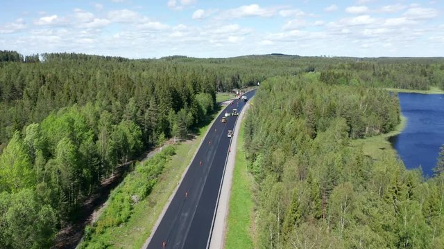 Drone footage slowly flying towards a group of road renovation machines. Pine forest on both side of the road and a small lake of the right hand side of the road. Filmed in realtime at 4k.