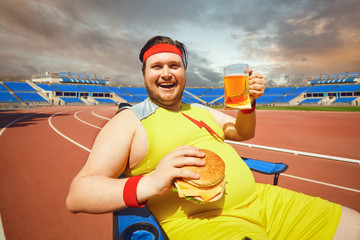 Fat man eating a burger and beer in training at the stadium.