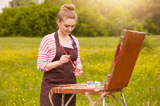 Portrait of beautiful female looking at watercolour samples on convas, mixed colours, has concentrated facial expression, woman wears casual clothes, has fair hair and bunch. Art and creation concept.