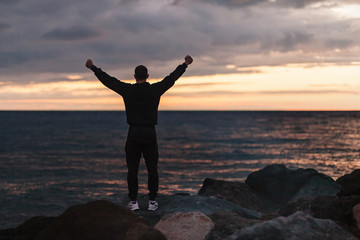 Rear view of a happy unidentified male athlete rejoices in a fulfilled dream by raising his hands up and looking at sunset by the sea on warm summer evening. Concept of achieving goals and results