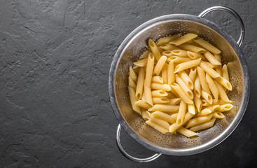 cooked Penne rigate pasta in stainless steel colander on Dark grey black slate background with copy space. top view