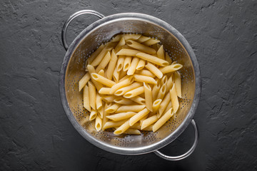 cooked Penne rigate pasta in stainless steel colander on Dark grey black slate background with copy space. top view