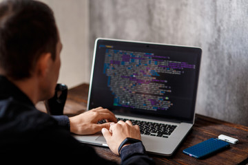 a male programmer in a black shirt is working at a laptop, close-up of the program code and developer's hands on a laptop. modern young programmer sitting at a wooden table at a computer