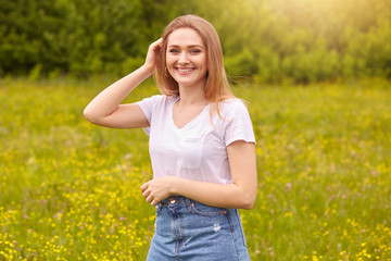Naklejka na ściany i meble Outdoor shot of woman posing in open air, young blonde model standing on grass in meadow, looking with romantic facial expression directly at camera, wearing white casual t shirt and denim skirt.