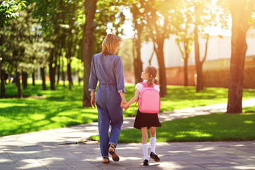 Fototapeta premium Parent and pupil of primary school go hand in hand. Woman and girl with pink backpack behind the back. Beginning of lessons. First day of fall