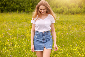 Naklejka na ściany i meble Ourdoor shot of young beautiful Caucasian girl in white t shirt and blue denim skirt posing against sunset in meadow, looking down, attractive female looking down. Lifestyle and happyness concept.