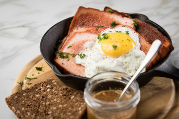Gebräunter Bayrischer Fleischkäse mit gebratenen Spiegelei in Gusseisenpfanne mit süßen Senf und Brot auf Holzbrett