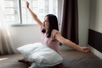 Portrait of Asian Woman is Stretching Her Hands on The Bedroom After Wake Up, Healthy Woman is Waking Up Morning Time on Bed in Restroom. Healthcare and Lifestyles Concept.