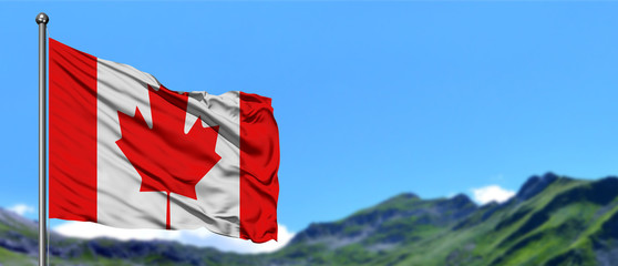 Canada flag waving in the blue sky with green fields at mountain peak background. Nature theme.