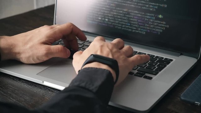 a male programmer in a black shirt is working at a laptop, close-up of the program code and developer's hands on a laptop. modern young programmer sitting at a wooden table at a computer