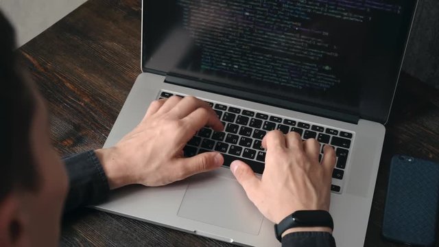 a male programmer in a black shirt is working at a laptop, close-up of the program code and developer's hands on a laptop. modern young programmer sitting at a wooden table at a computer