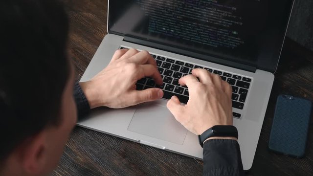 a male programmer in a black shirt is working at a laptop, close-up of the program code and developer's hands on a laptop. modern young programmer sitting at a wooden table at a computer