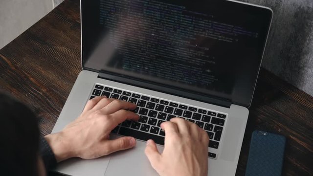 a male programmer in a black shirt is working at a laptop, close-up of the program code and developer's hands on a laptop. modern young programmer sitting at a wooden table at a computer