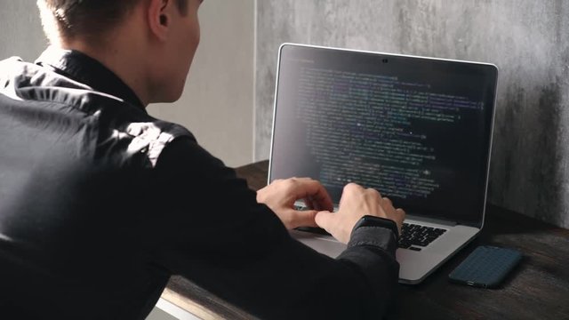 a male programmer in a black shirt is working at a laptop, close-up of the program code and developer's hands on a laptop. modern young programmer sitting at a wooden table at a computer