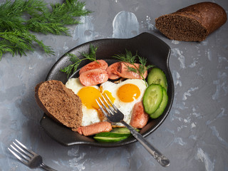 Breakfast of scrambled eggs, sausages and dill for breakfast in a black ceramic plate. Slices and a slice of bread on a gray background.