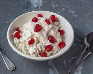 A plate of homemade cottage cheese with sour cream and dried cherries for a light breakfast. Teaspoons lie side by side on a gray background. Shot from close range.