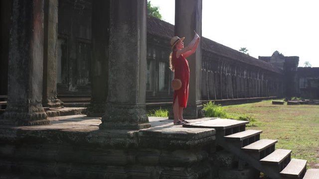 Sliding view of young woman coming on the porch of Angkor Wat temple and taking picture on her smartphone. The temple was built in 12th century in Cambodia and dedicated to Vishnu. Cambodia