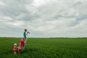 Obraz na płótnie Canvas Funny children in bright celebratory clothes play on green field with wheat. Pink suitcase, folding ladder, megaphone. Boys are happy together on vacation in village..Unity with nature