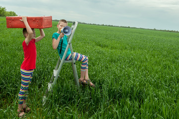 Funny children in bright celebratory clothes play on green field with wheat. Pink suitcase, folding ladder, megaphone. Boys are happy together on vacation in village..Unity with nature