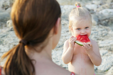 Happy blonde girl with her mum eating a slice of watermelon, candid outdoor lifestyle portrait