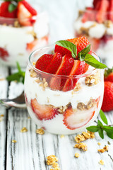 Healthy breakfast of strawberry parfaits made with fresh fruit, yogurt and granola over a rustic white table. Shallow depth of field with selective focus on glass jar in front. Blurred background.