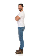 Fototapeta premium Smiling Man In Jeans And White T-shirt Is Standing With Arms Crossed And Looking At Camera. Side View.