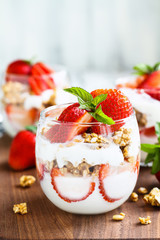 Healthy breakfast of strawberry parfaits made with fresh fruit, yogurt and granola over a rustic wood table. Shallow depth of field with selective focus on glass jar in front. Blurred background.